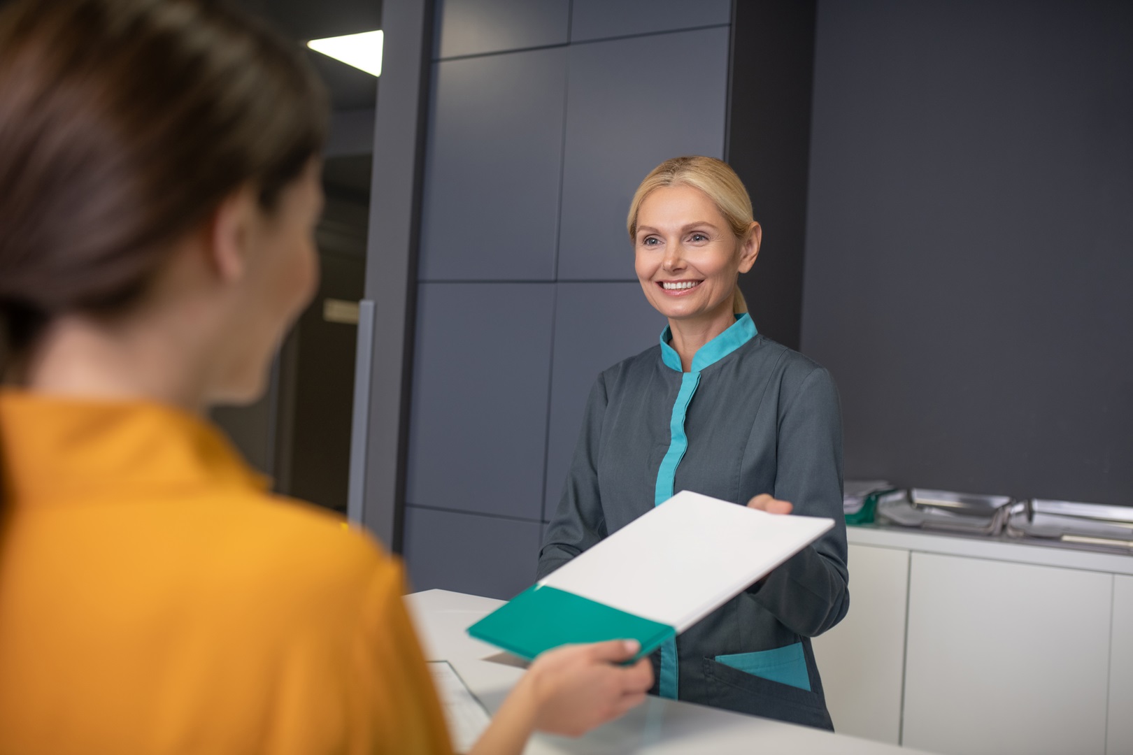 Blond woman in grey jacket talking to new visitor Receptionist helping with patient documentation by greeting patients