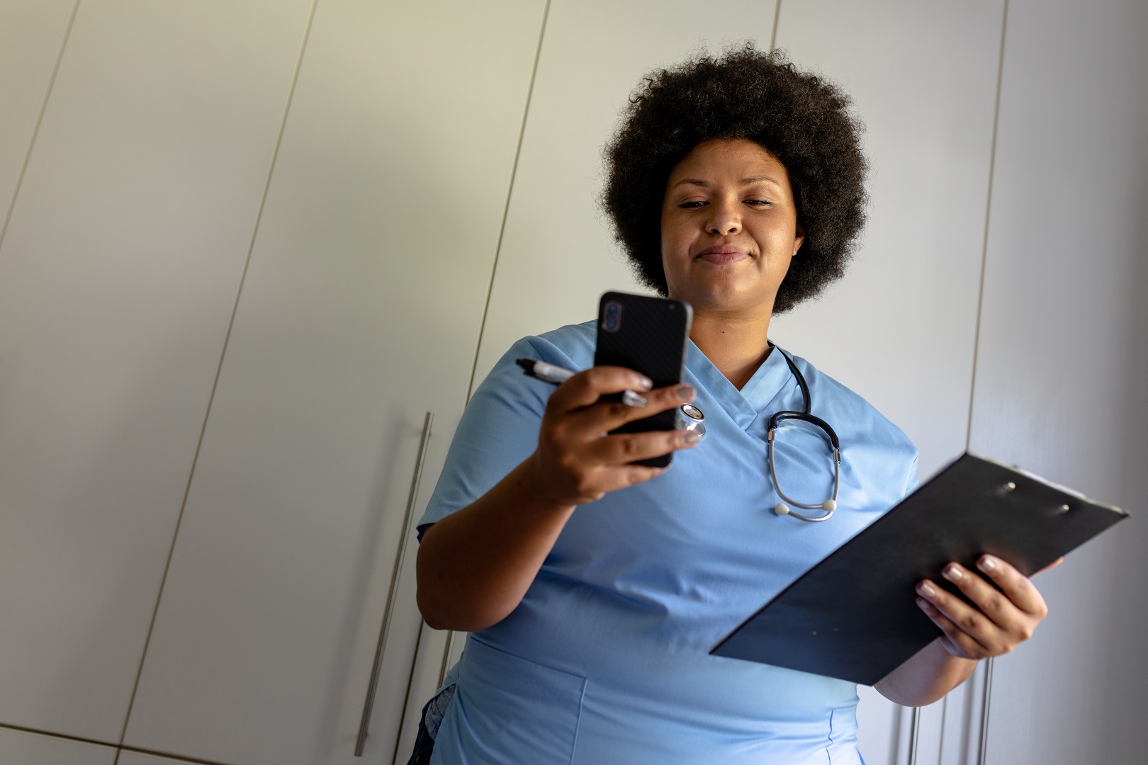 African american mid adult female nurse using smart phone while holding clipboard in hospital. unaltered, healthcare worker, wireless technology, expertise and occupation concept. Medical receptionist sending patient reminder through phone text
