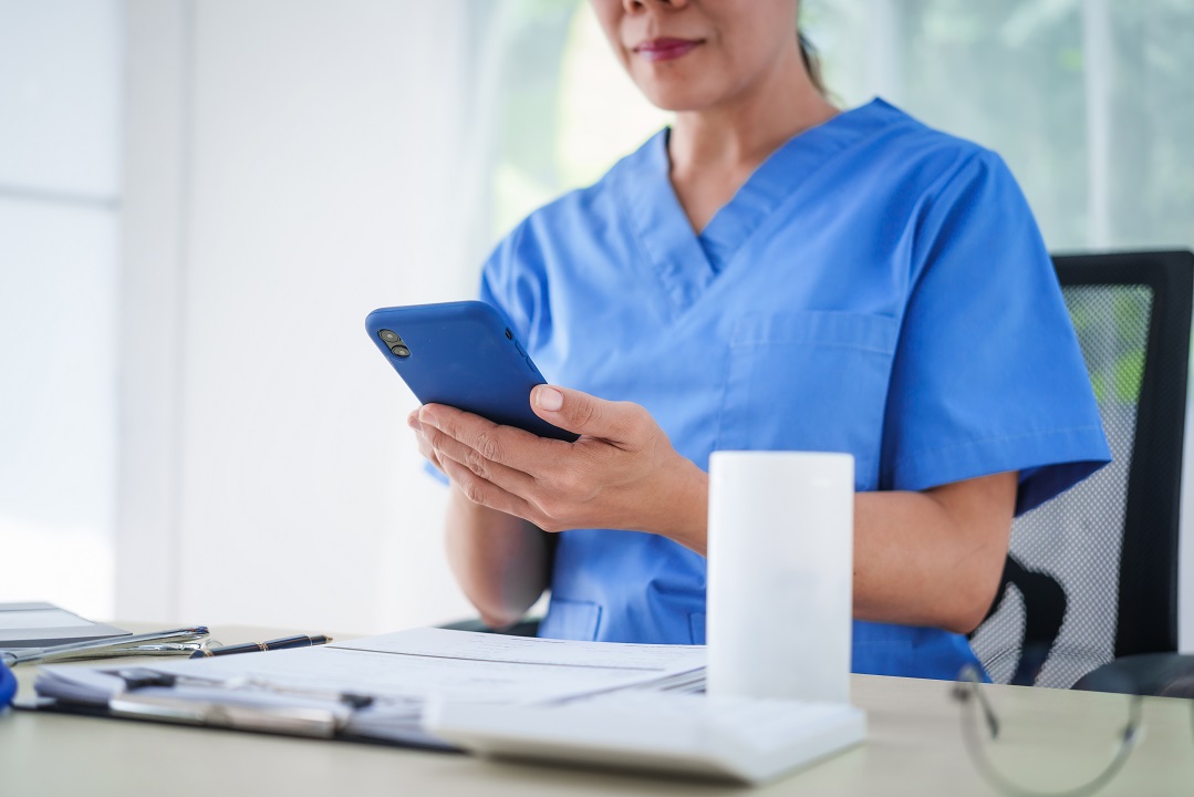 An Asian female nurse sits at a table, talking on the phone with Physical therapist sending patient appointment reminders via phone text