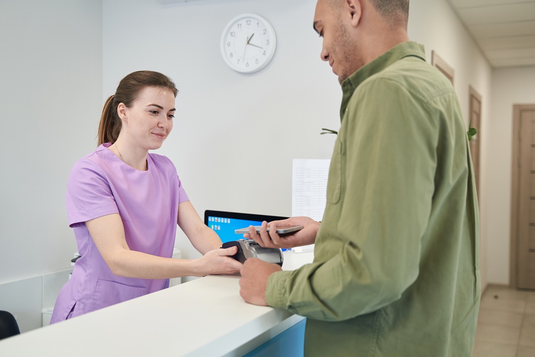 Male client paying for clinic services with smartphone Receptionist facilitating patient collections with patient via card terminal